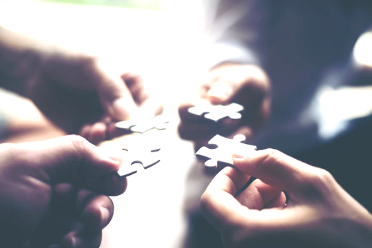 Four hands holding puzzle pieces, connecting in the center, symbolizing partnership and collaboration.